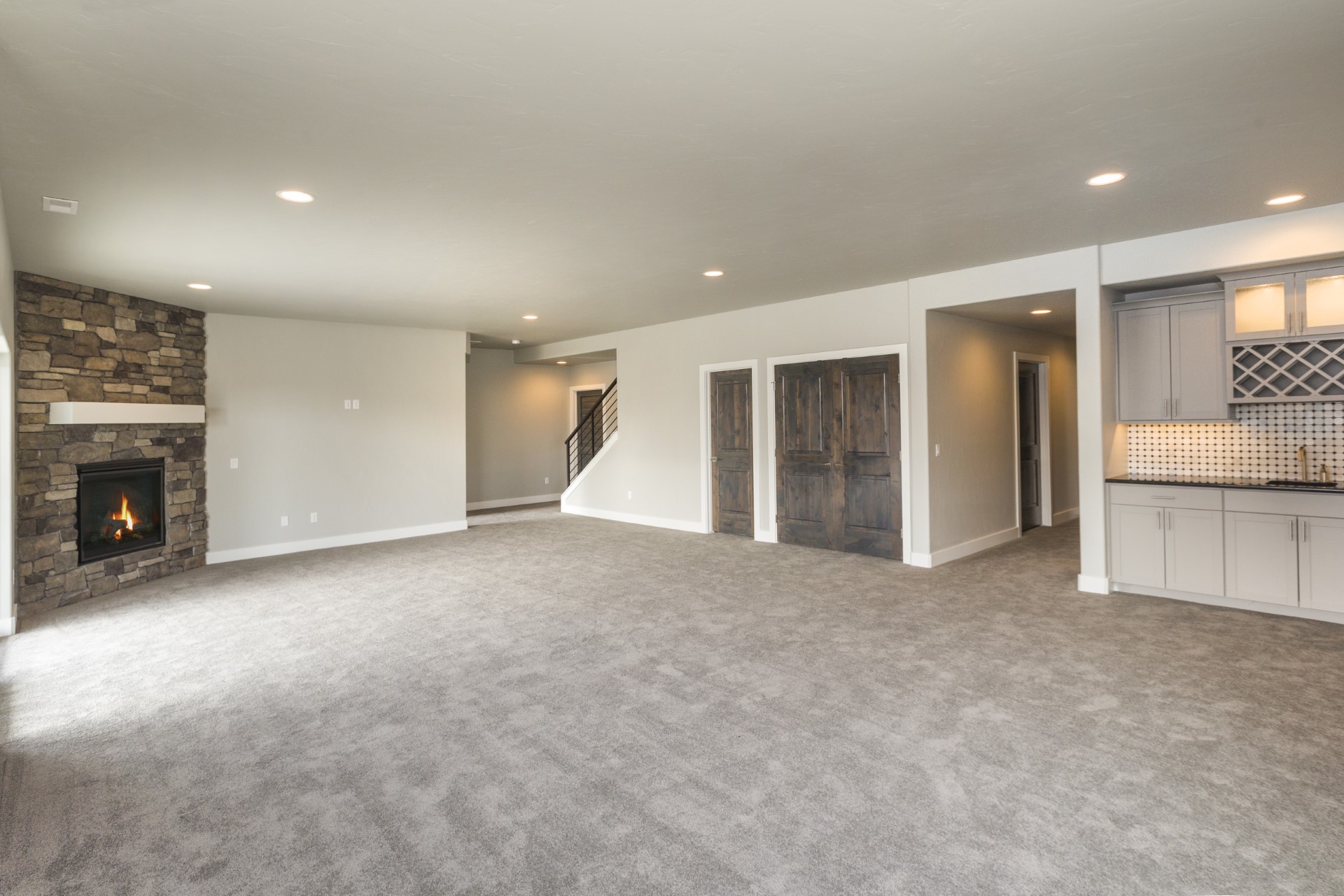 Completed basement bonus room with light-colored carpeting, grey walls, a large bar on the right for dining, and a stone fireplace on the left with a lit fire.